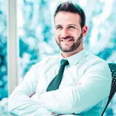 Uomo sorridente con camicia bianca e cravatta, braccia conserte, seduto alla scrivania. Il suo sguardo è rivolto verso l'esterno, come un back office manager che pianifica il futuro.