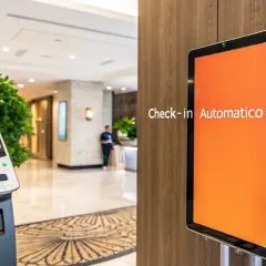 Un totem per il **smart check-in** in un hotel moderno. Un tablet mostra "Check-in Automatico" su sfondo arancione.