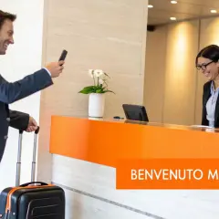 Uomo con valigia fa check-in express, sorridendo alla receptionist. Un benvenuto memorabile.