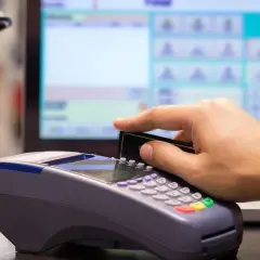 Pagamento con carta su POS, con registratore di cassa sullo sfondo.