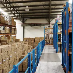 Magazzino pieno di scatoloni e scaffalature blu. Si prepara la dismissione cespiti, con merci in attesa di essere catalogate o spostate.