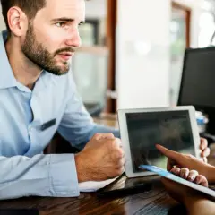 Un impiegato dell'hotel mostra a una cliente cosa fare in caso di overbooking, usando un tablet per trovare soluzioni.