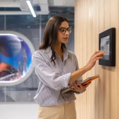 Donna interagisce con un pannello touch, parte dell'automazione hotel, tenendo un tablet.
