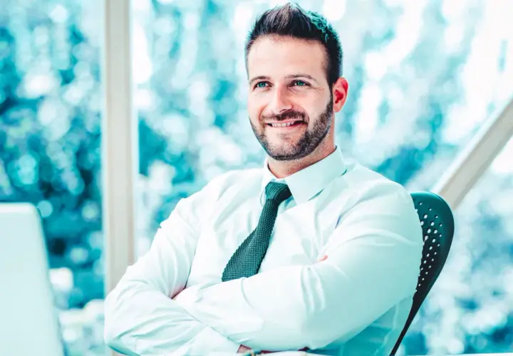 Uomo sorridente con camicia bianca e cravatta, braccia conserte, seduto alla scrivania. Il suo sguardo è rivolto verso l'esterno, come un back office manager che pianifica il futuro.