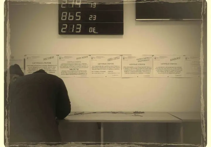 Un uomo si china su un tavolo, mentre sullo sfondo un display digitale mostra numeri. Informazioni su "codice univoco cos'è" e altri servizi sono affisse ai muri.