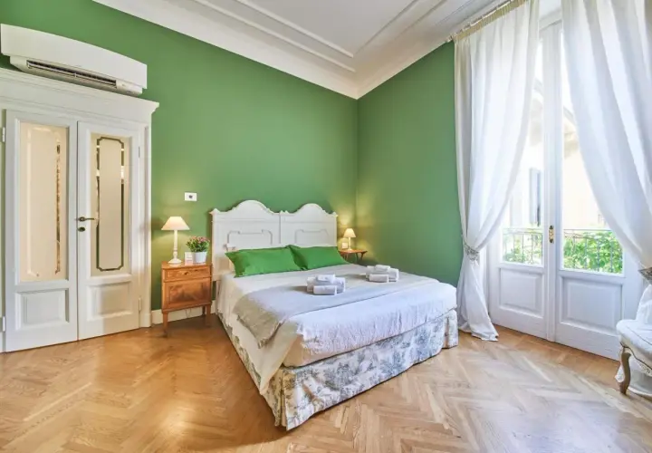 Camera da letto accogliente con pareti verdi, letto bianco, tende bianche e parquet. Ideale per capire la differenza tra foresteria e B&B.