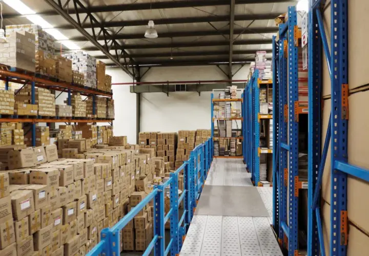 Magazzino pieno di scatoloni e scaffalature blu. Si prepara la dismissione cespiti, con merci in attesa di essere catalogate o spostate.