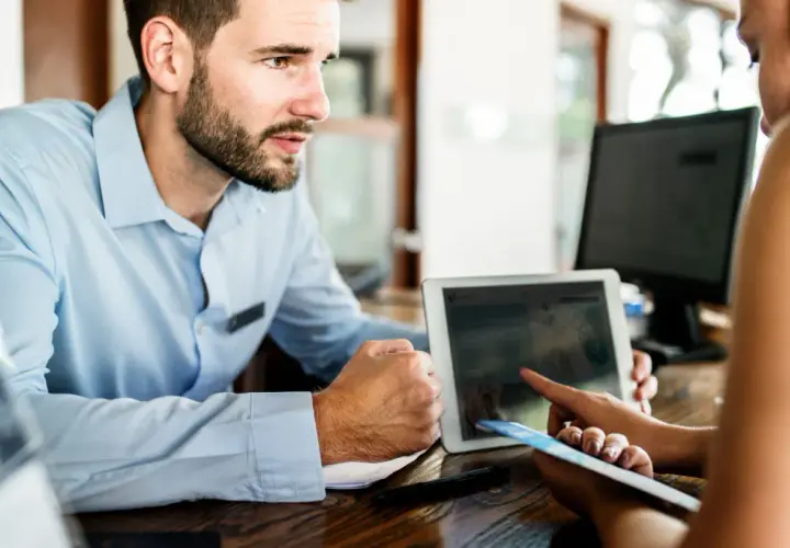 Un impiegato dell'hotel mostra a una cliente cosa fare in caso di overbooking, usando un tablet per trovare soluzioni.