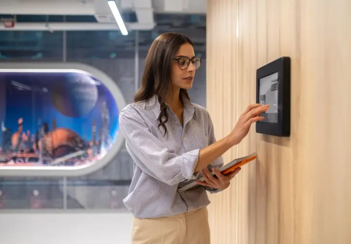 Donna interagisce con un pannello touch, parte dell'automazione hotel, tenendo un tablet.