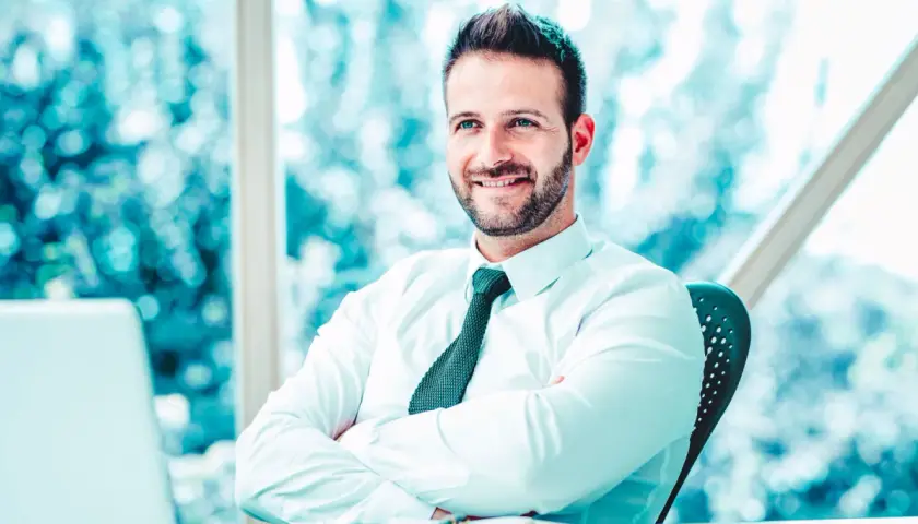 Uomo sorridente con camicia bianca e cravatta, braccia conserte, seduto alla scrivania. Il suo sguardo è rivolto verso l'esterno, come un back office manager che pianifica il futuro.