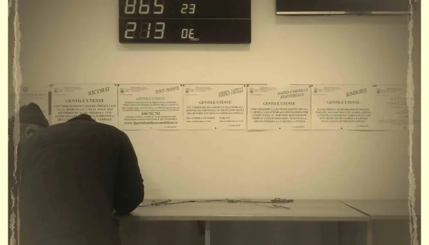Un uomo si china su un tavolo, mentre sullo sfondo un display digitale mostra numeri. Informazioni su "codice univoco cos'è" e altri servizi sono affisse ai muri.
