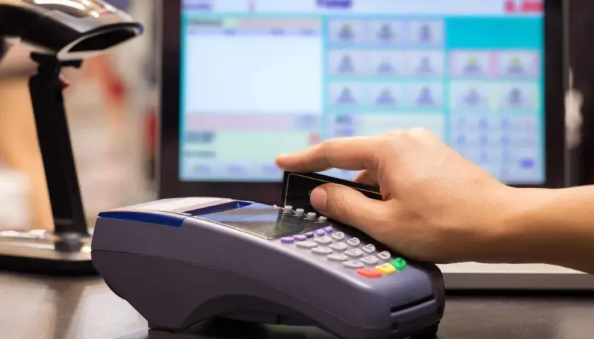 Pagamento con carta su POS, con registratore di cassa sullo sfondo.