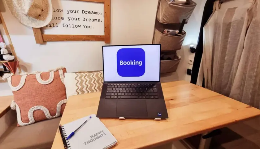 Laptop con logo Booking aperto su un tavolo di legno, accanto a un quaderno "Happy Thoughts". Pianifica il tuo prossimo viaggio con credito booking.