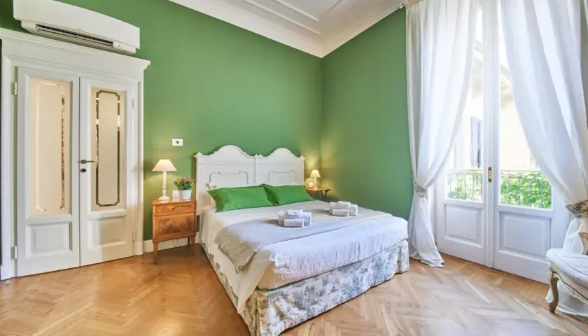 Camera da letto accogliente con pareti verdi, letto bianco, tende bianche e parquet. Ideale per capire la differenza tra foresteria e B&B.