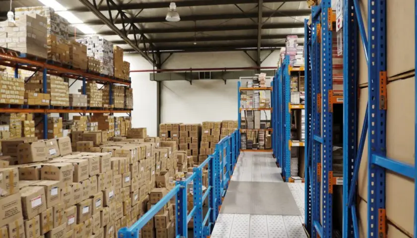 Magazzino pieno di scatoloni e scaffalature blu. Si prepara la dismissione cespiti, con merci in attesa di essere catalogate o spostate.