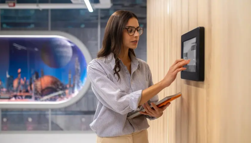 Donna interagisce con un pannello touch, parte dell'automazione hotel, tenendo un tablet.