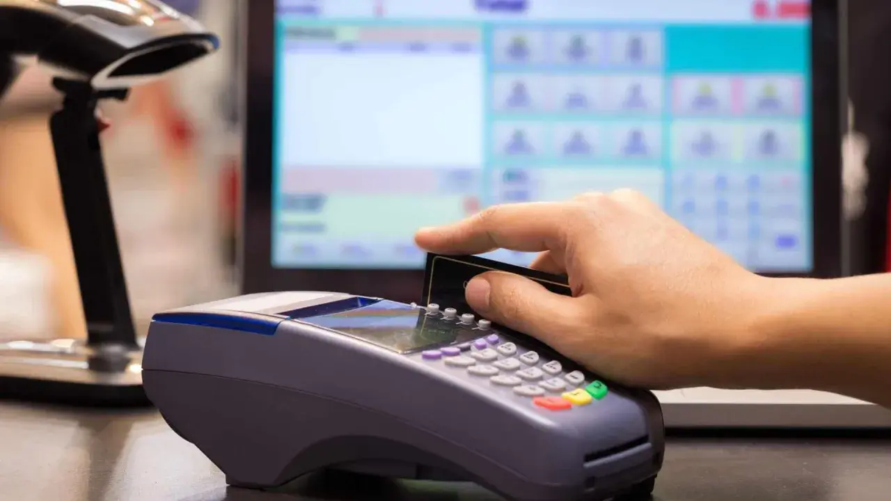 Pagamento con carta su POS, con registratore di cassa sullo sfondo.