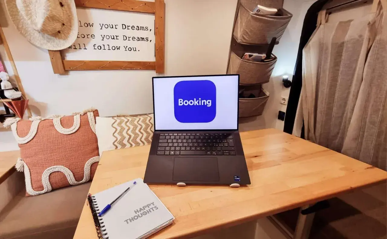 Laptop con logo Booking aperto su un tavolo di legno, accanto a un quaderno "Happy Thoughts". Pianifica il tuo prossimo viaggio con credito booking.