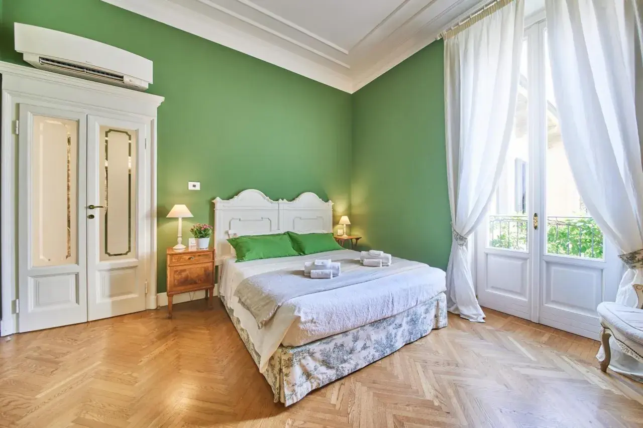 Camera da letto accogliente con pareti verdi, letto bianco, tende bianche e parquet. Ideale per capire la differenza tra foresteria e B&B.