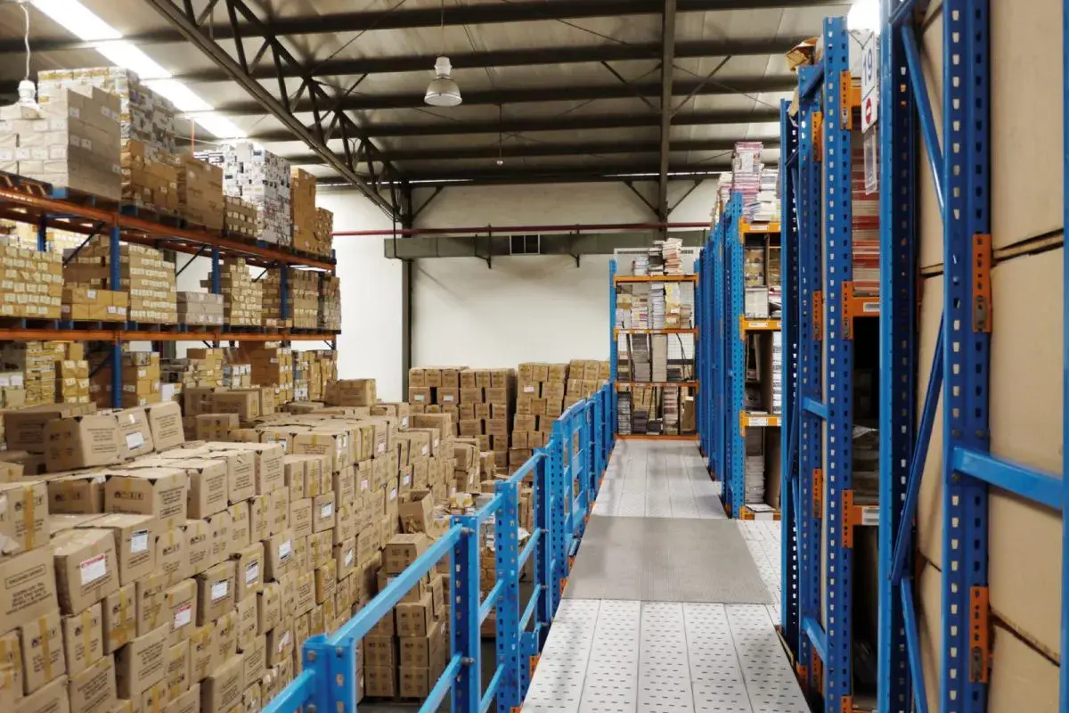Magazzino pieno di scatoloni e scaffalature blu. Si prepara la dismissione cespiti, con merci in attesa di essere catalogate o spostate.