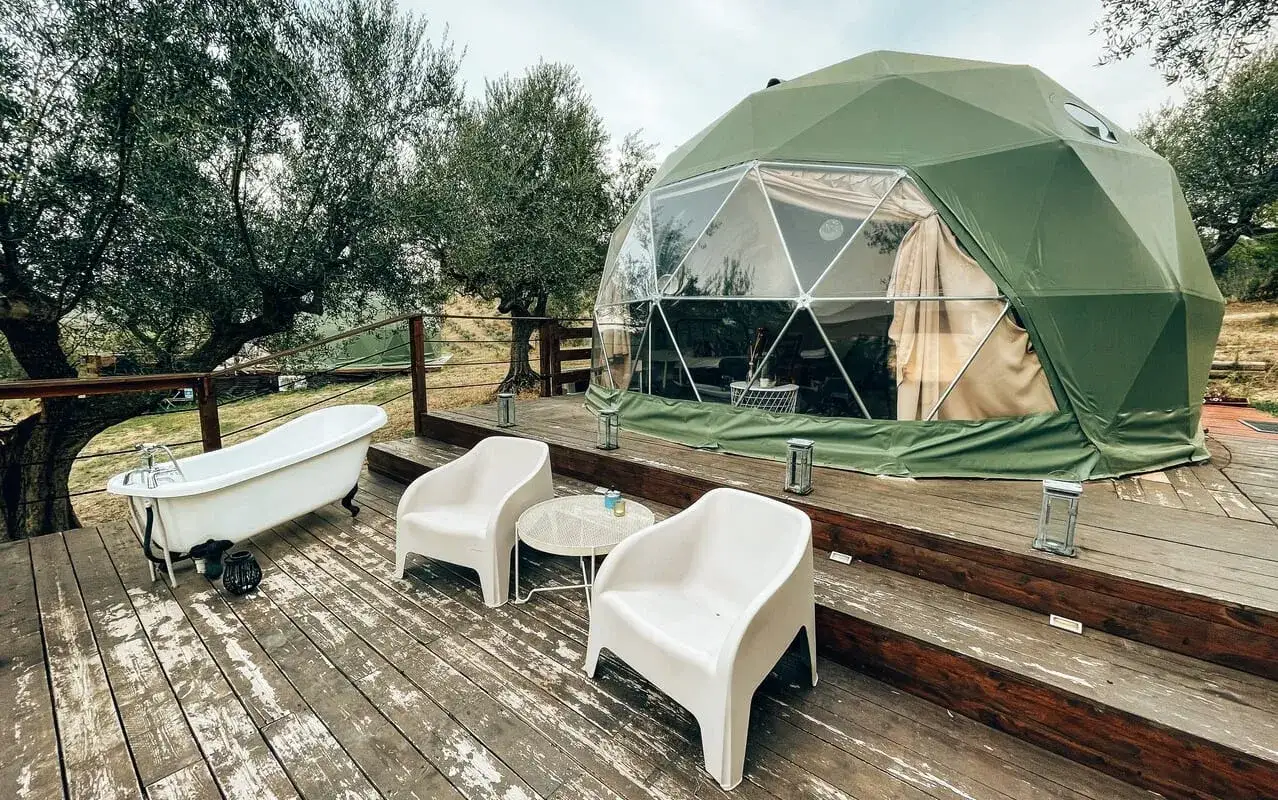 La tenda geodetica verde, un esempio di glamping significato, offre un bagno all'aperto e sedie comode su una terrazza di legno tra gli ulivi.