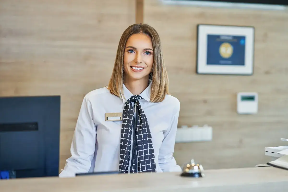 Sorriso accogliente di una receptionist, simbolo di eccellente customer care. Dietro il bancone, un computer e un campanello per servire.