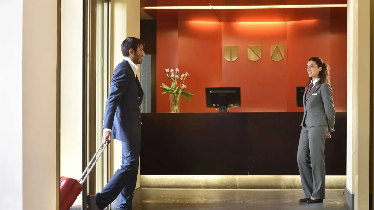 Uomo con valigia rossa si avvicina alla reception per il check-in in hotel. La receptionist sorride.
